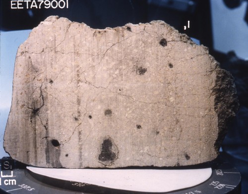 Marsmeteorit EETA79001 in Querschnitt. Dieser Meteorit wurde 1979 in Antarktika in der Region Viktorialand gefunden. Er besteht aus Basalt-Gestein mit dunklen Stellen aus ehemals geschmolzenem Glas, welches Spuren von Gasen der Marsatmosphäre enthält [46]. Der Würfel links unten ist 1 cm3 groß.( Urheber: NASA, USA. Public domain)